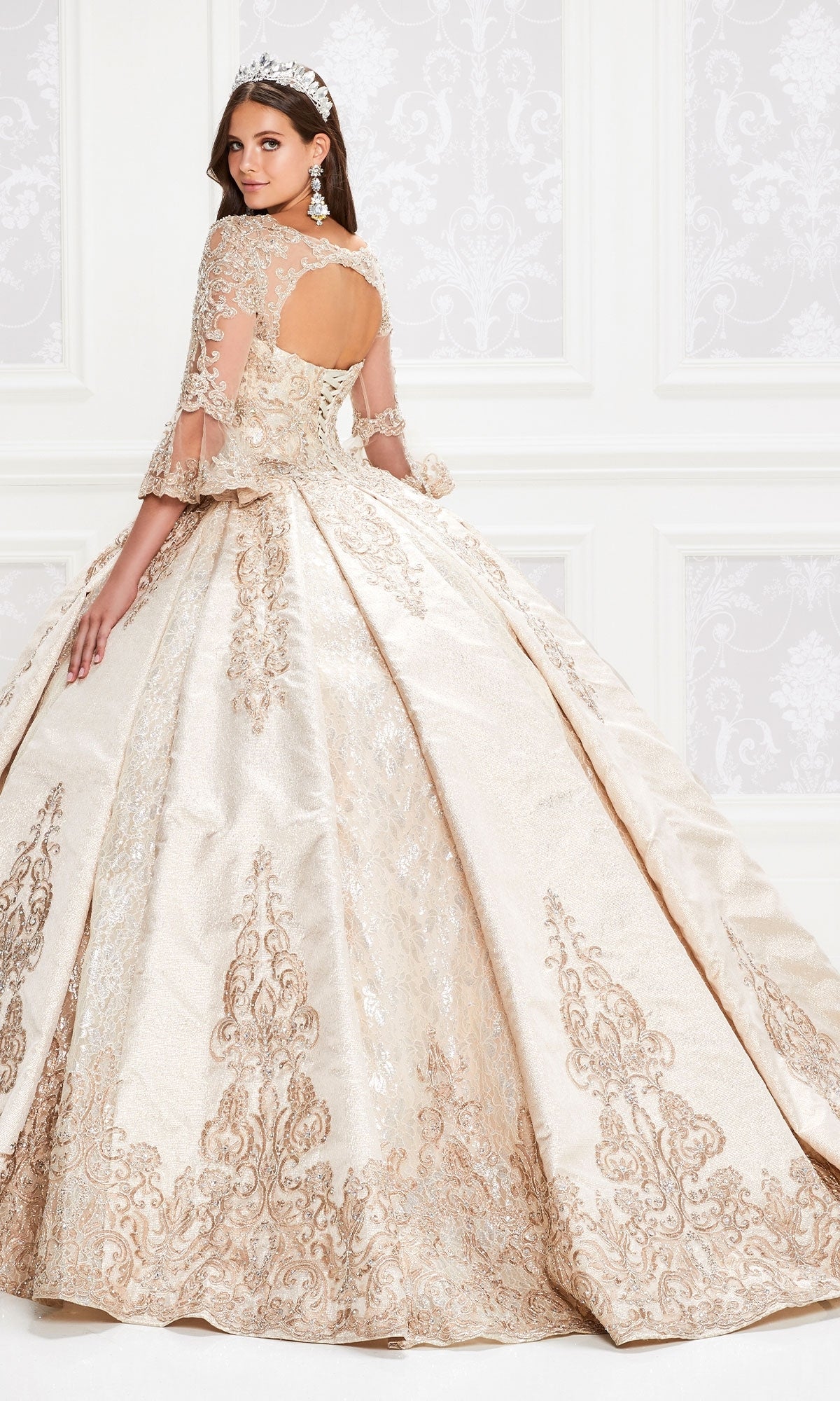 A young woman wears the Princesa PR12004 Quinceañera Dress with elegant gold and cream tones, lace bell sleeves, a tiara, and statement earrings as she stands in an ornate room facing slightly backward.